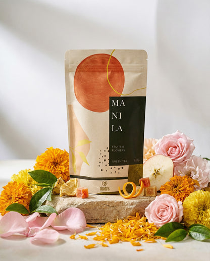 Alanda's Tea MANILA Doypack Produktfoto mit Rosen, Ringelblumen, Apfel und Ananas auf Steinpodest