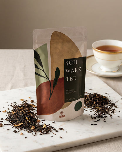 SCHWARZTEE Ceylon & Darjeeling von Alandas – Doypack mit losem schwarzen Tee und Teetasse auf Marmorfläche